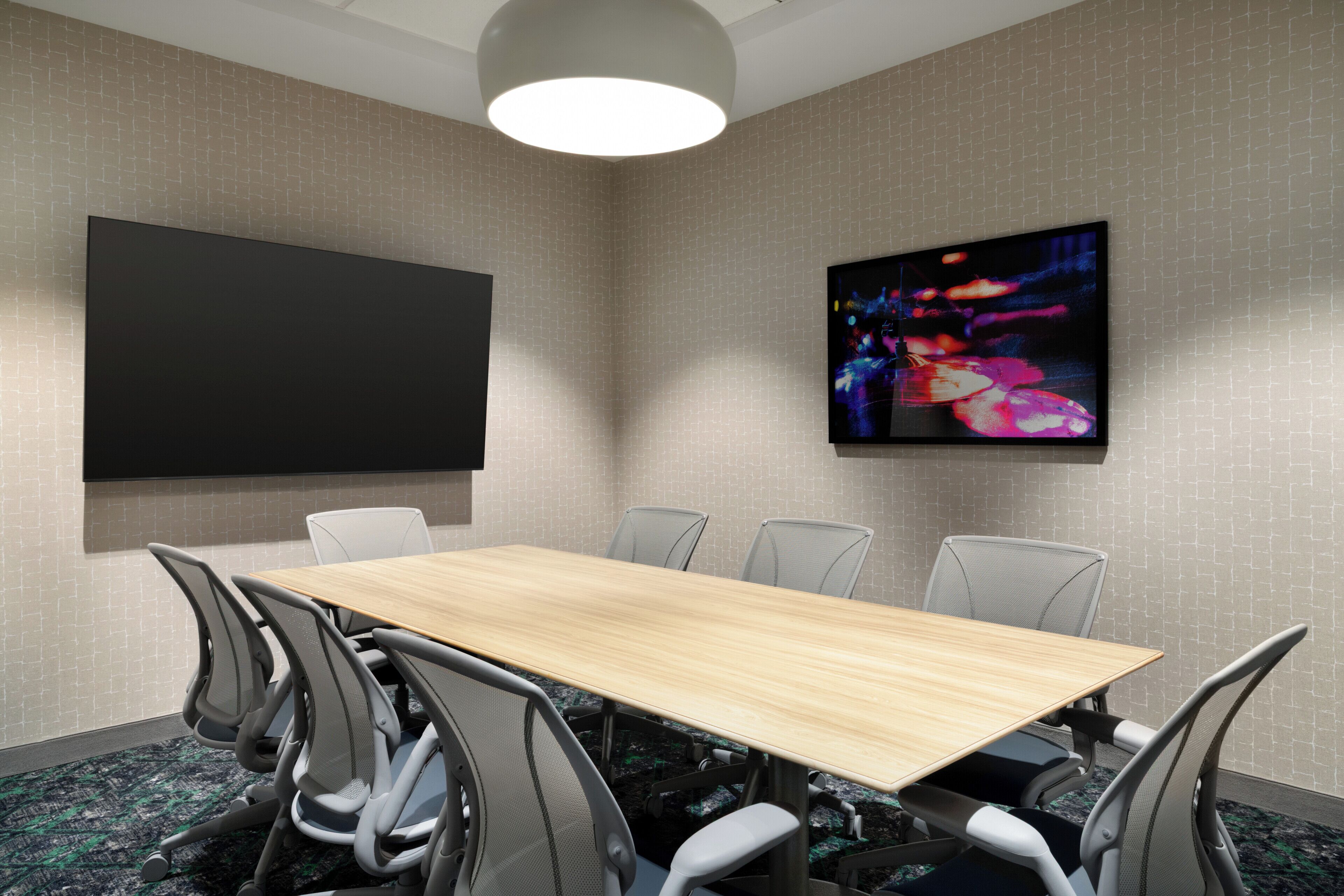 A conference room with a long table, six chairs, a large flat-screen TV, and a colorful abstract painting on the wall.