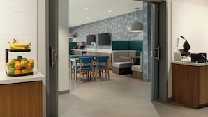 A modern kitchen with a dining area, featuring a white countertop with fruits, a wooden cabinet, and a seating arrangement with blue chairs. - Home2 Suites By Hilton Denton (Denton)
