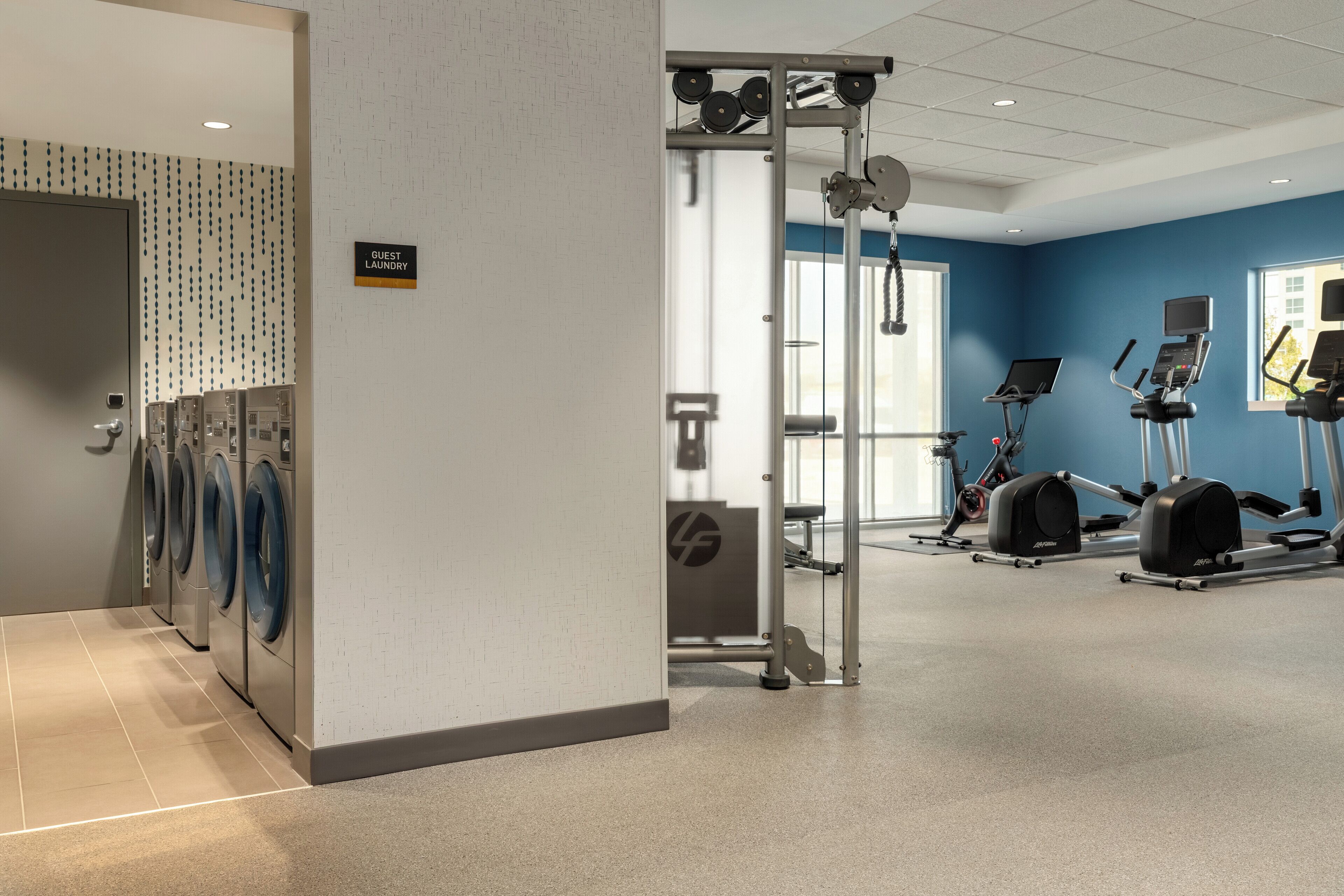 A hotel gym with a wall-mounted laundry area, exercise equipment, and a sign indicating a guest laundry room.