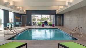 An indoor swimming pool with clear glass walls, lounge chairs, and a view of the outside. - Home2 Suites By Hilton Denton (Denton)
