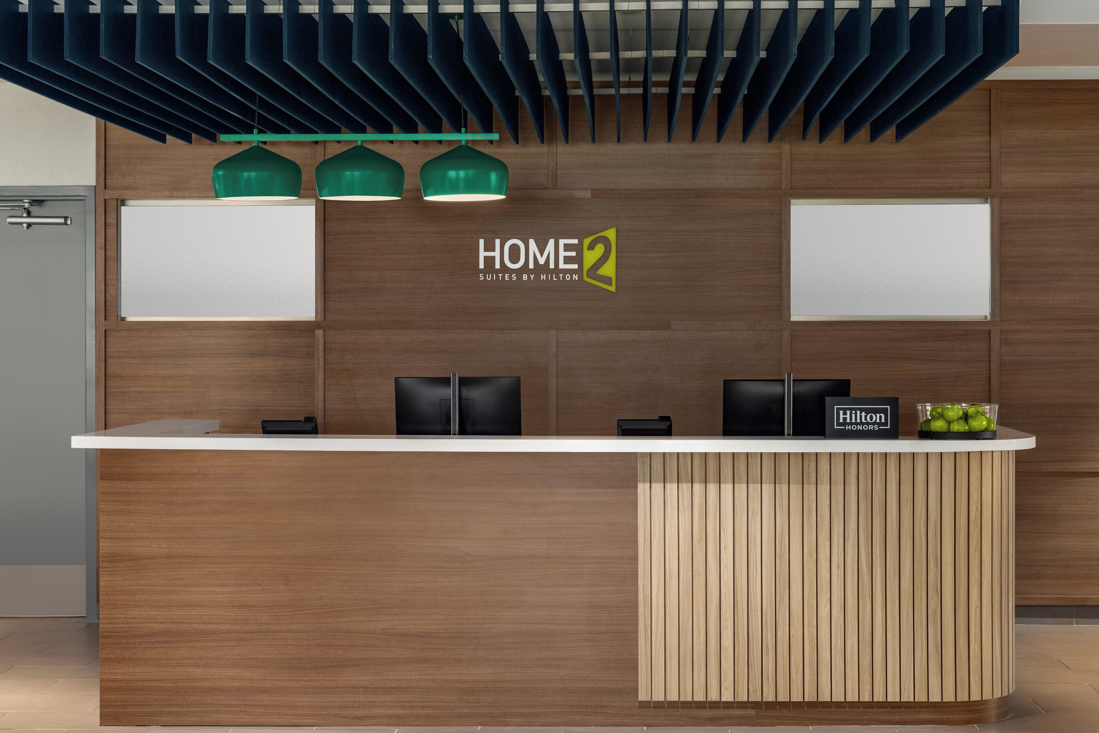 A modern hotel reception desk with a wooden panel design, green pendant lights, and a sign reading "HOME2 Suites by Hilton".