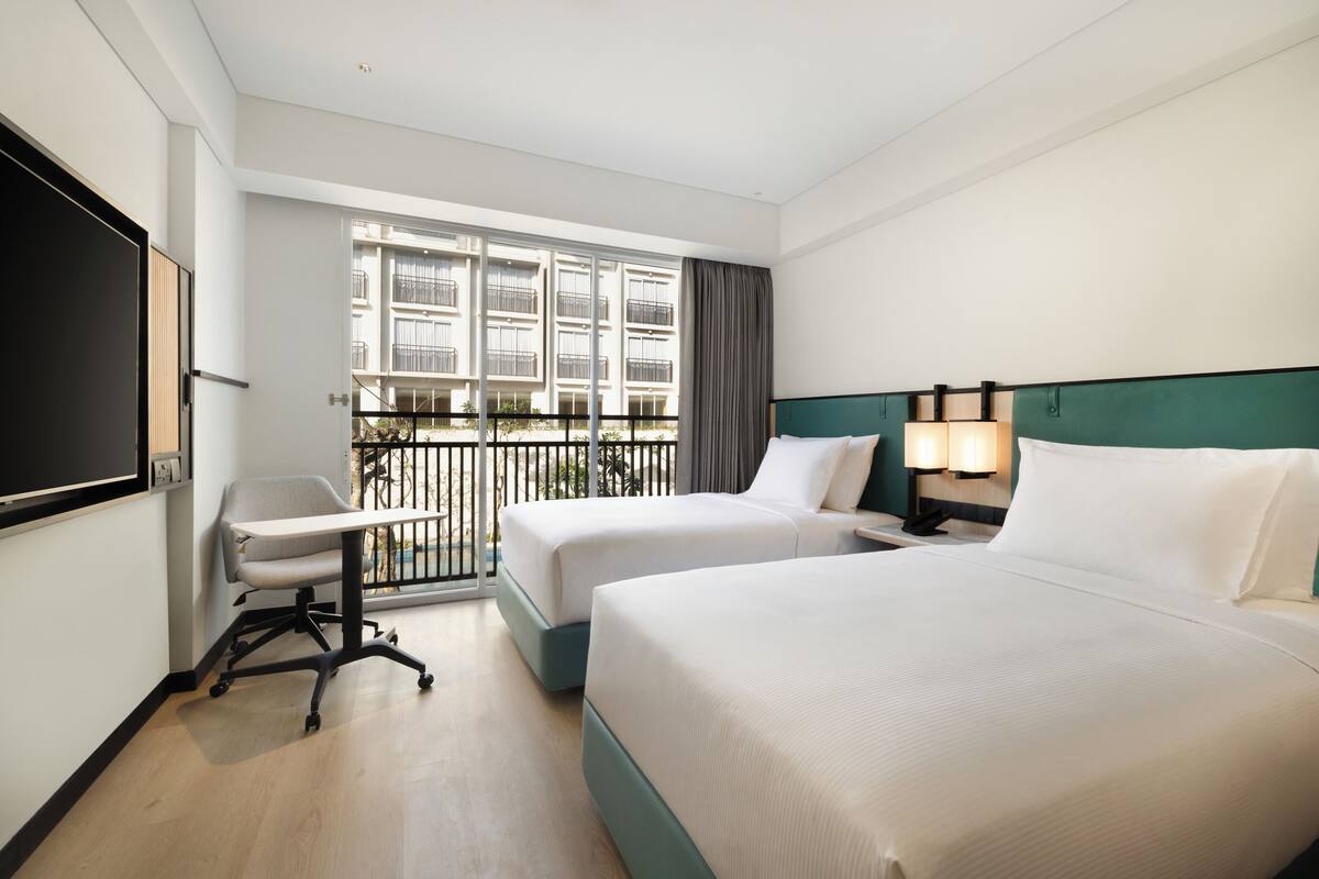 A hotel room with two beds, a desk, and a large window with a balcony.