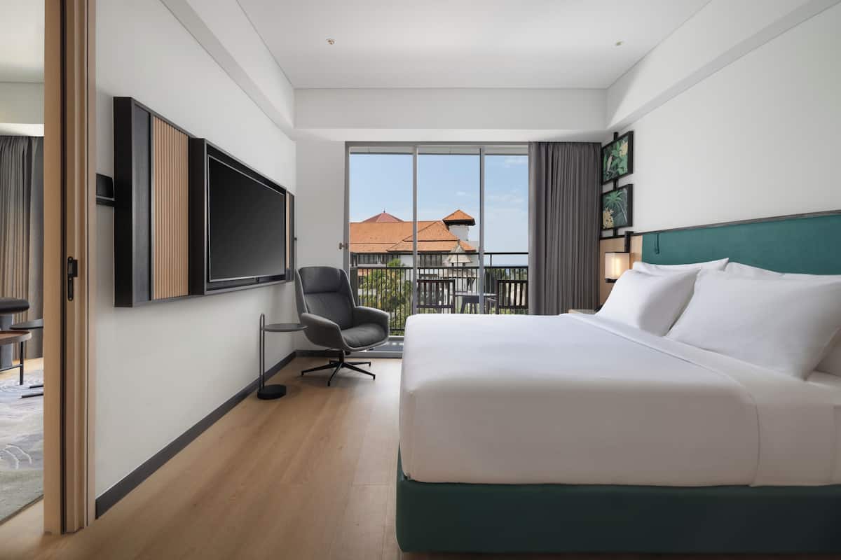 A hotel room with a large bed, a TV mounted on the wall, a chair, and a balcony with a view of buildings.
