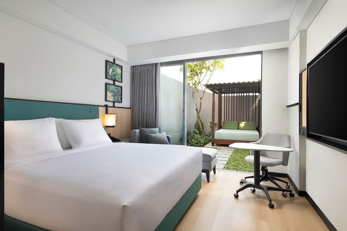 A modern hotel room with a large bed, a desk, a chair, and a small garden area visible through the sliding doors.