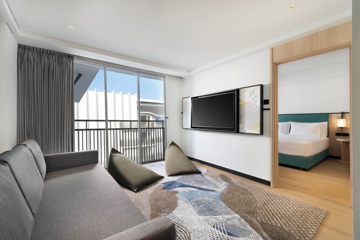 A modern hotel room with a large sofa, a flat-screen TV, a bed with a green headboard, and a balcony with curtains.