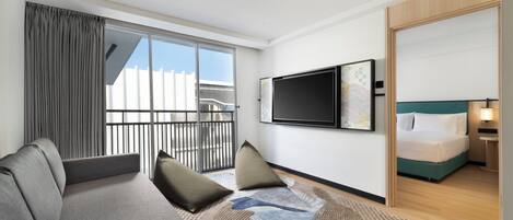 Una habitación de hotel moderna con un sofá grande, un televisor de pantalla plana, una cama con cabecera verde y un balcón con cortinas.