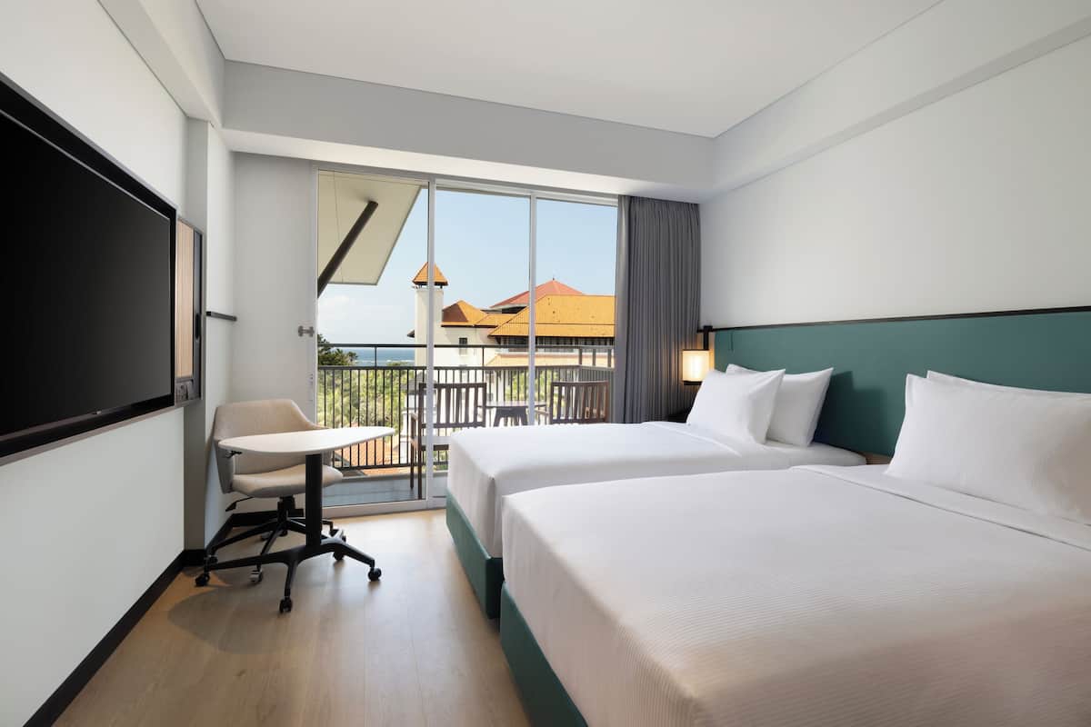 A hotel room with two beds, a desk, and a balcony with a view of buildings.