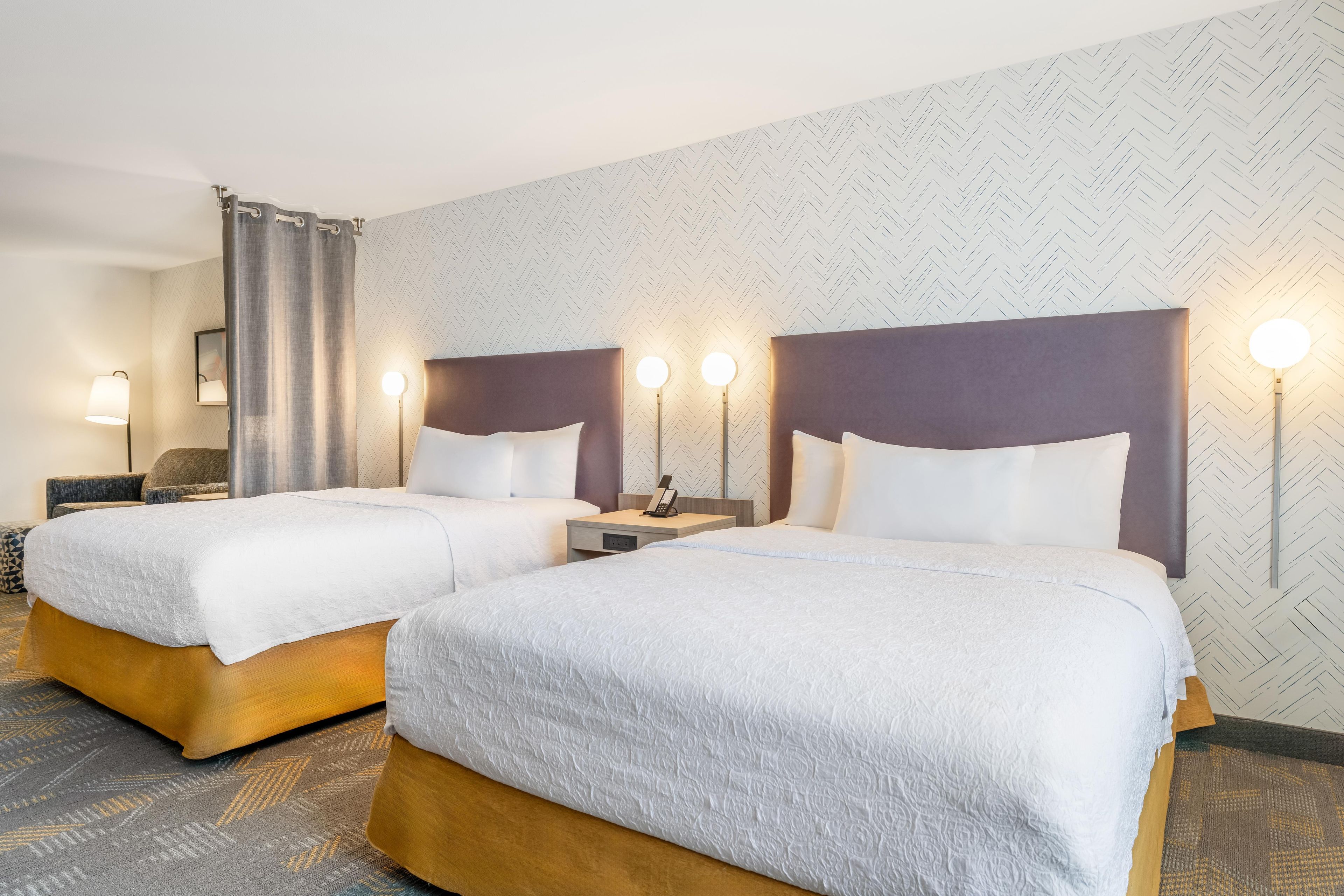 A hotel room with two beds, a desk, a chair, and a nightstand with a lamp.