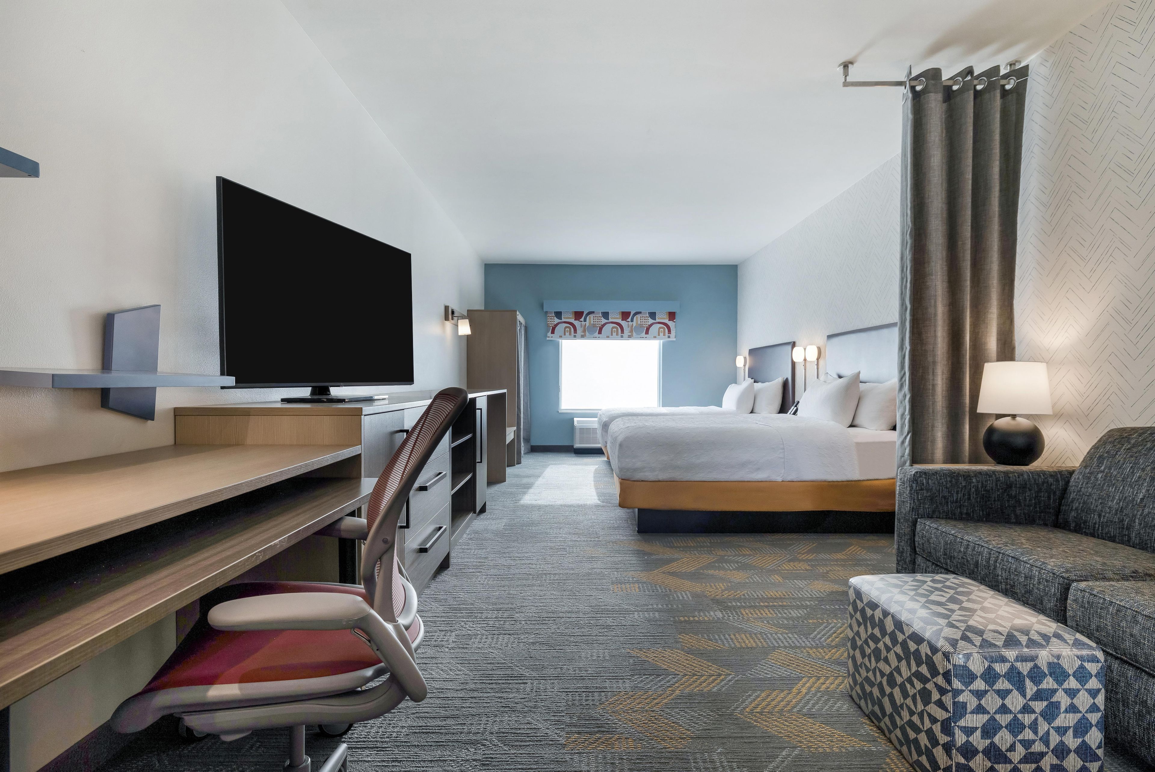 A hotel room with a bed, a desk with a flat-screen TV, a chair, a sofa, and a patterned carpet.