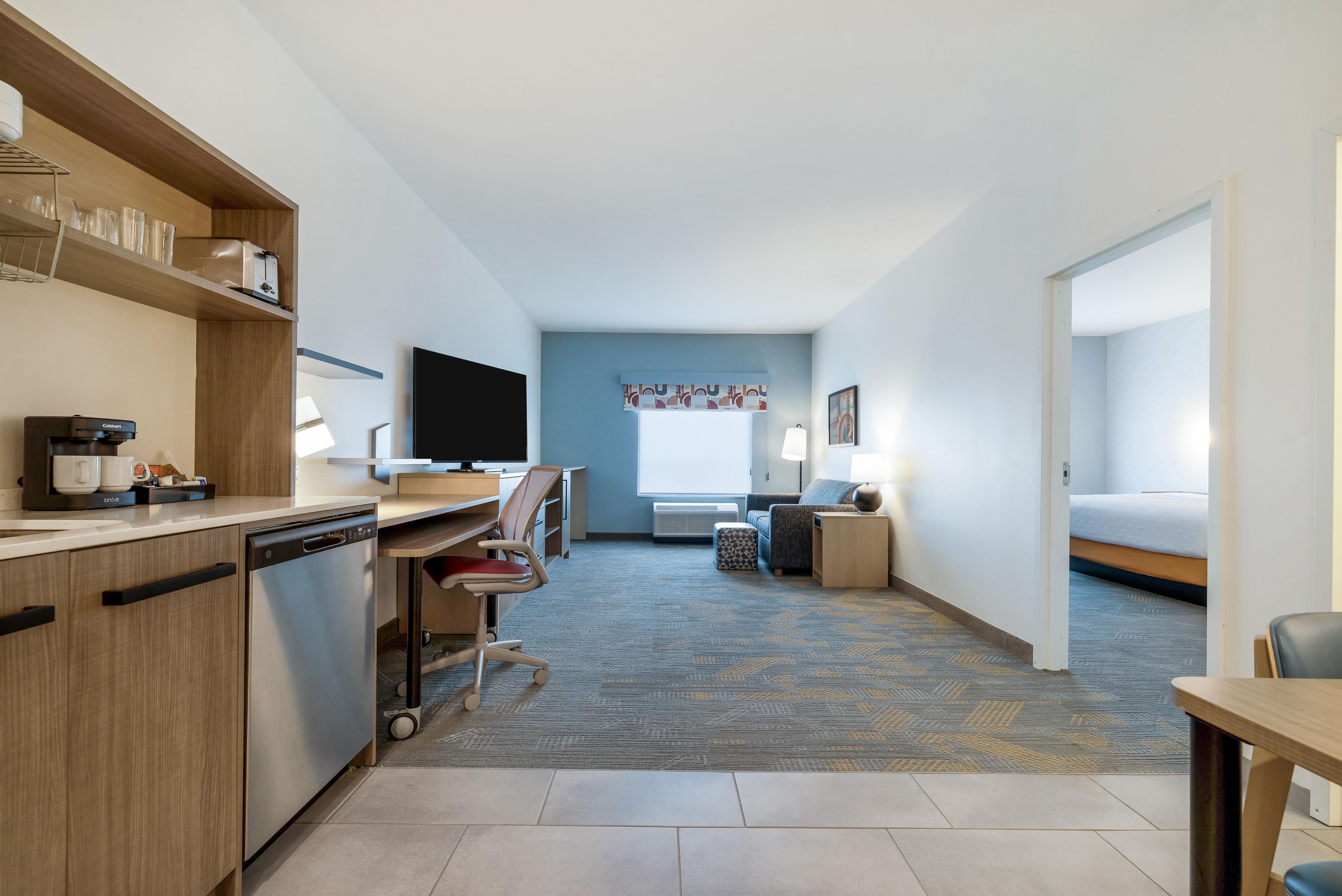 A hotel room with a kitchenette, a desk, a chair, a sofa, a TV, and a bed.