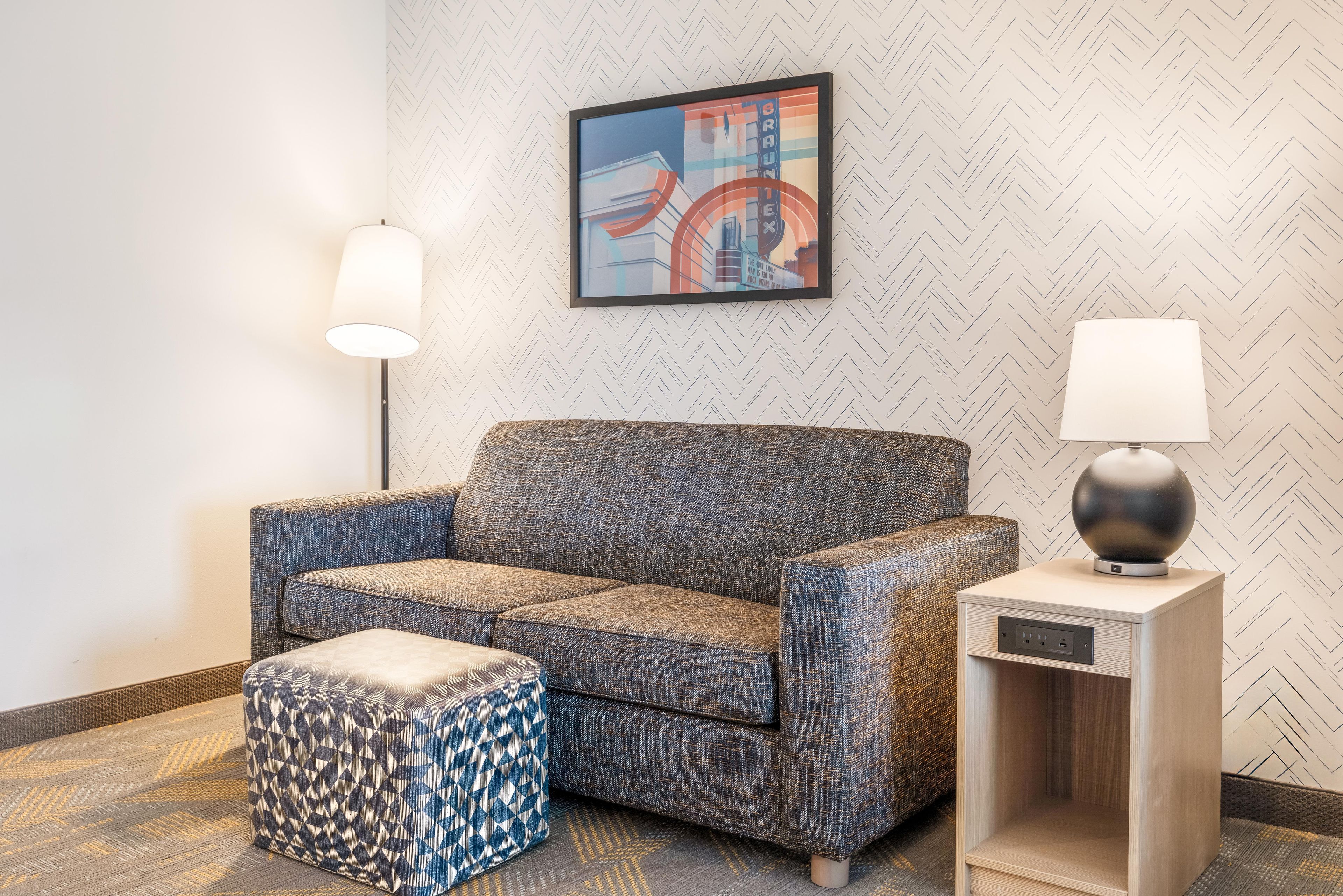 Un salon moderne avec un canapé à motifs, un pouf assorti, une table d’appoint avec une lampe et une œuvre d’art encadrée accrochée au mur.