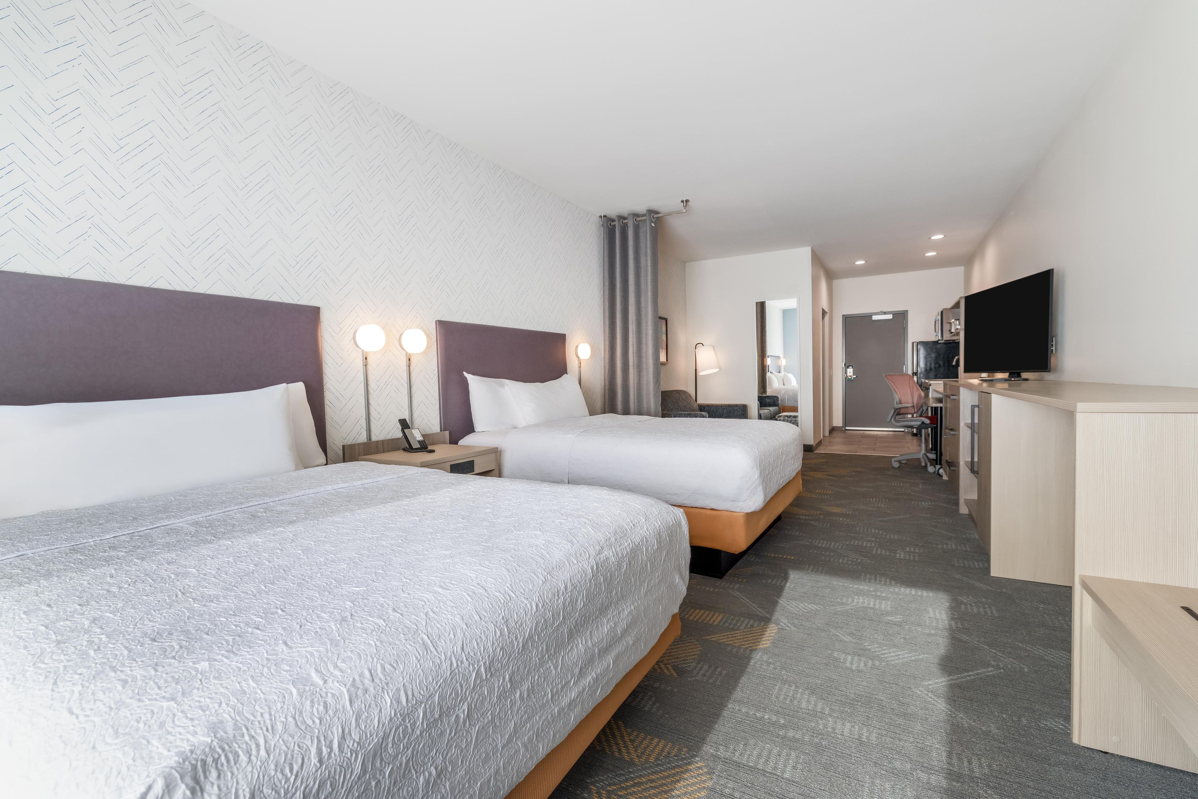 A hotel room with two beds, a desk, a television, and a bathroom in the background.