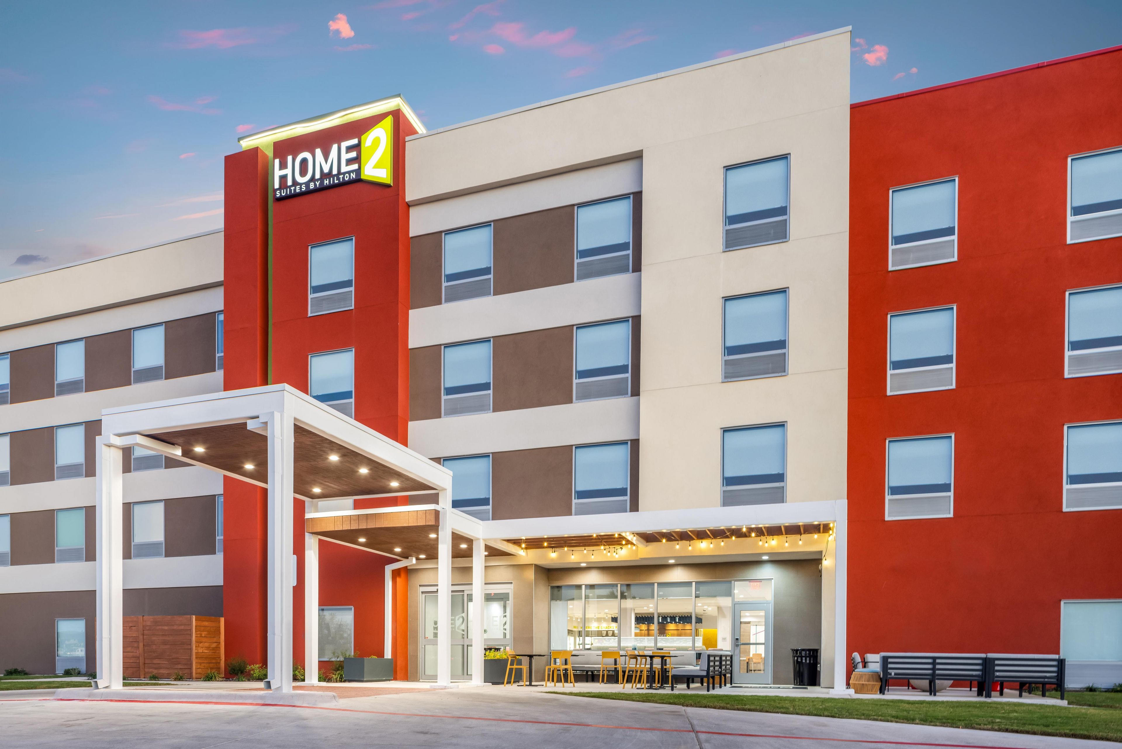 A modern hotel with a red and beige facade, multiple windows, and a covered entrance area with seating.