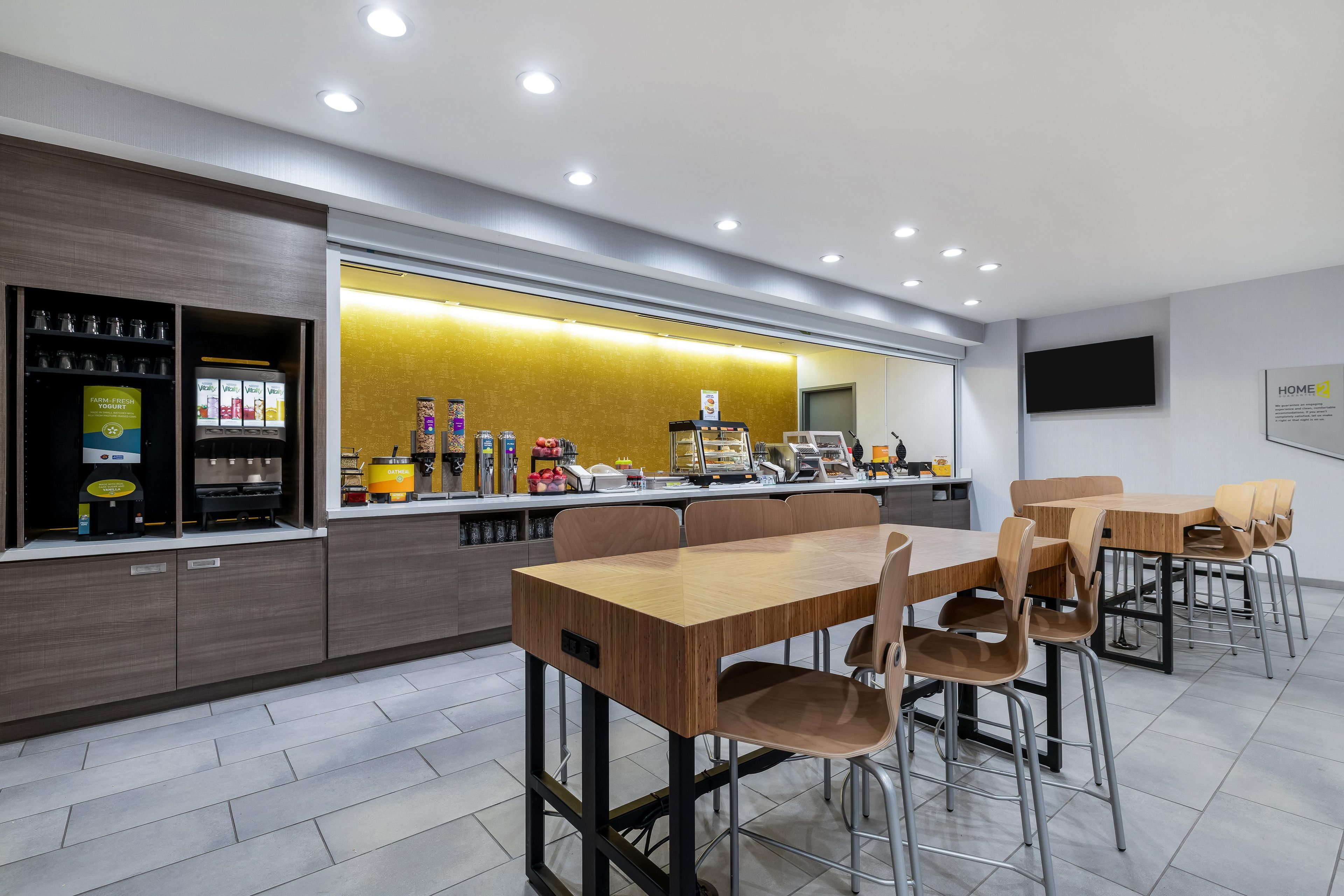 A modern cafe with a self-service counter, seating area, and a flat-screen TV on the wall.