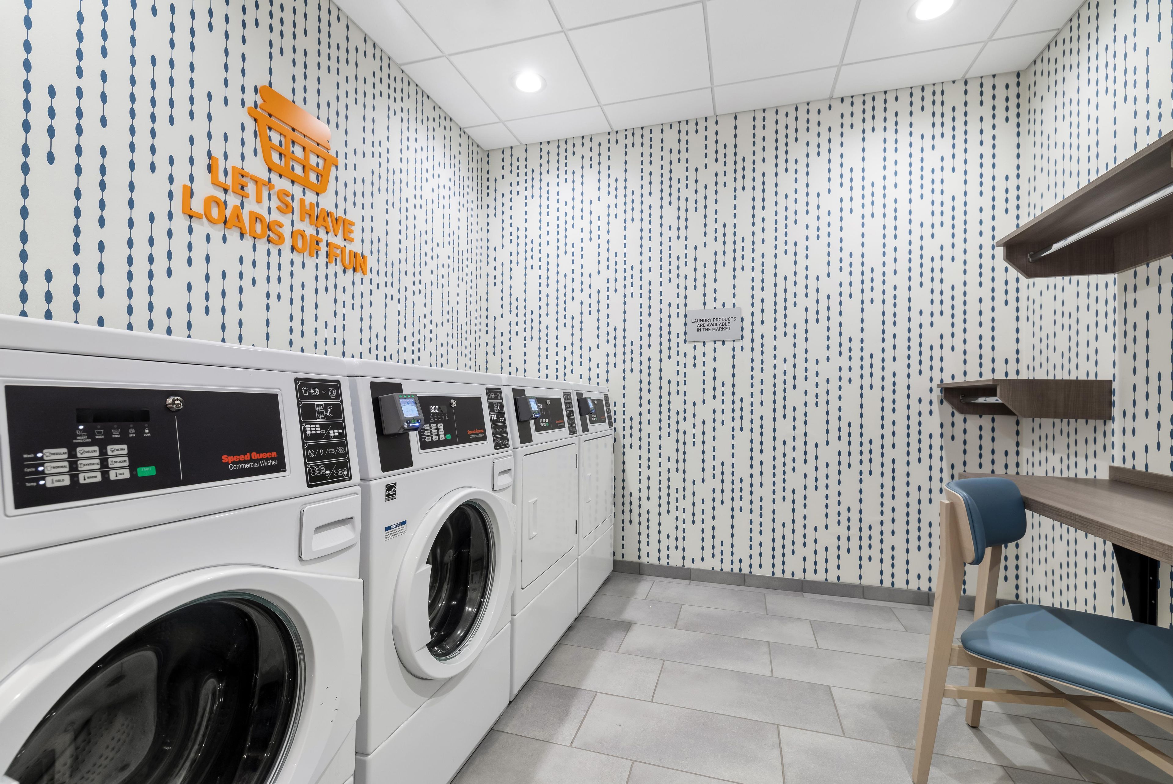 A laundry facility with multiple washing machines, a sign indicating laundry products are available, and a patterned wall.