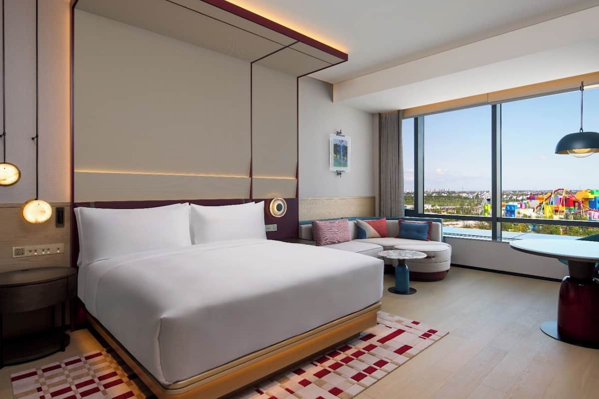 Une chambre d’hôtel moderne avec un grand lit, un canapé, une petite table et une vue sur un parc d’attractions aux couleurs vives.