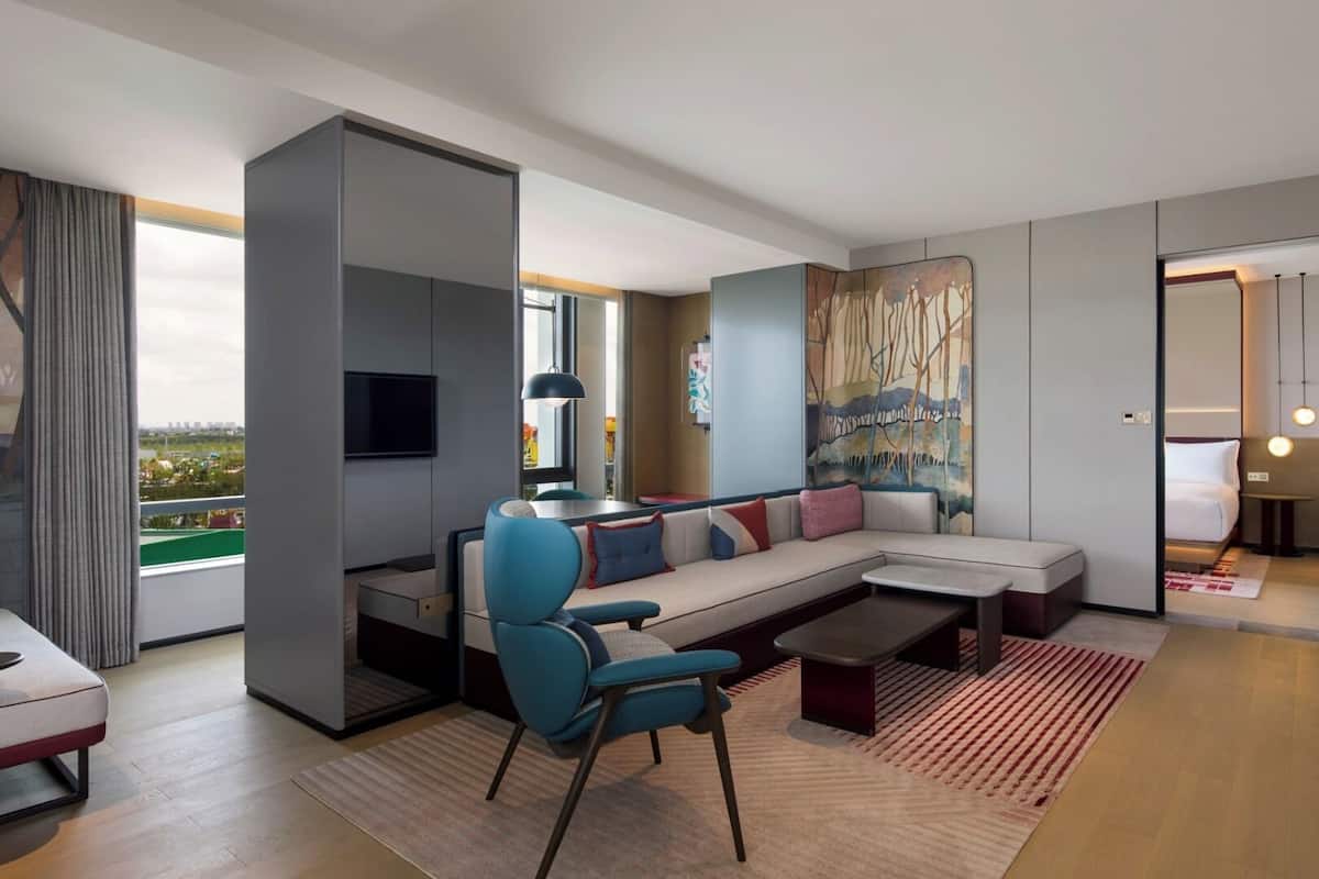 Une chambre d’hôtel moderne avec une grande fenêtre, un canapé, une table basse, un téléviseur et un tableau au mur.