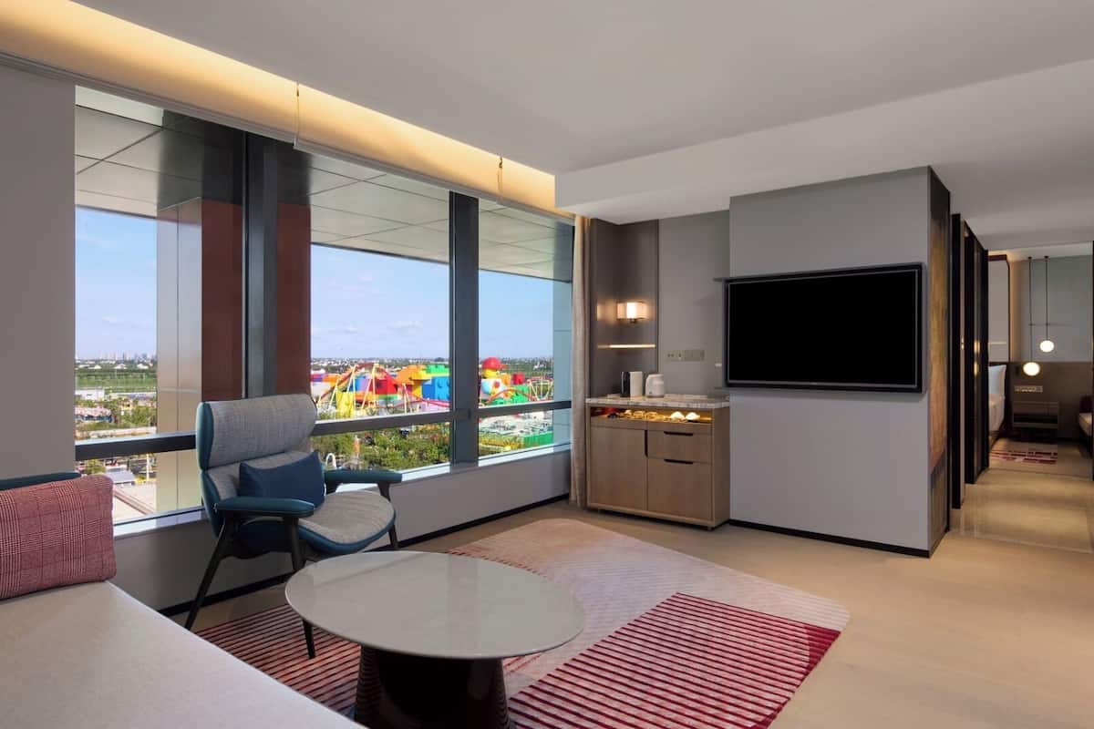 Une chambre d’hôtel moderne dotée d’une grande fenêtre, d’un téléviseur à écran plat, d’un coin salon avec une table et d’un coin cuisine.