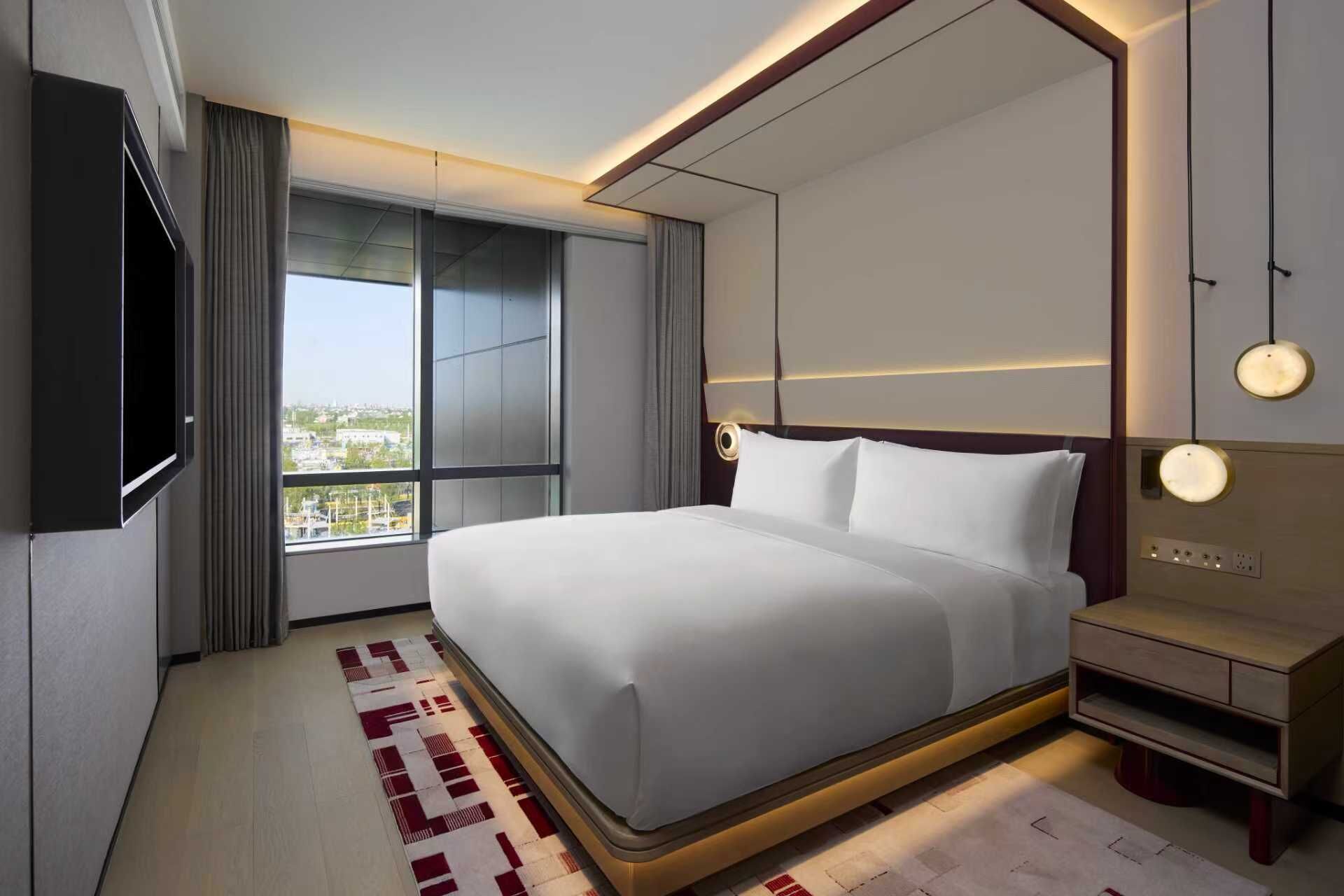 A modern hotel room with a large bed, a flat-screen TV, a bedside table, and a view of the city through large windows.