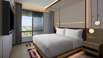 A modern hotel room with a large bed, a flat-screen TV, a bedside table, and a view of the city through large windows.