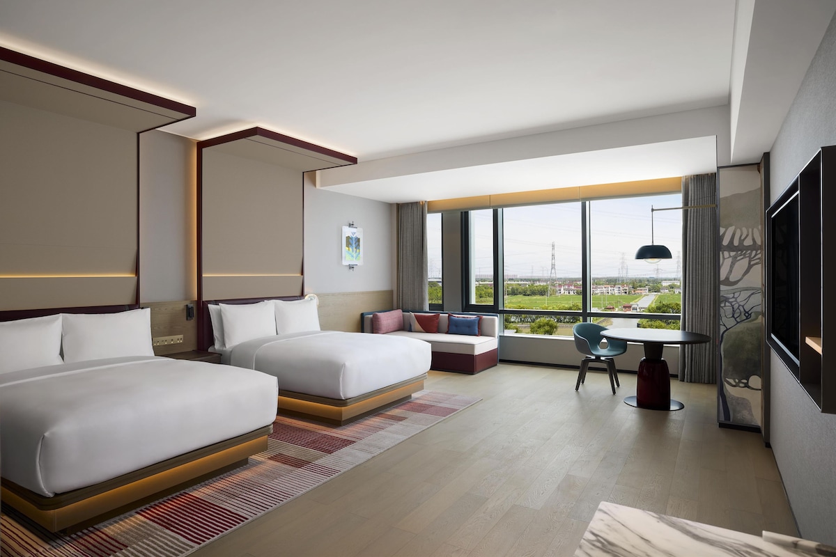 Une chambre d’hôtel moderne dotée de deux lits, d’un canap é, d’une table à manger et d’une grande fenêtre donnant sur la ville.