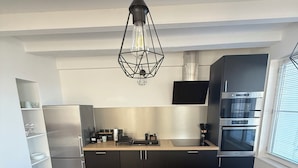 Una cocina moderna con armarios negros, un refrigerador de acero inoxidable, horno empotrado y una lámpara colgante.