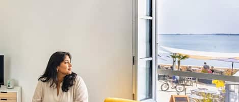 A room with a large window overlooking a beach, a laptop on a table, and a yellow cushion on a couch.