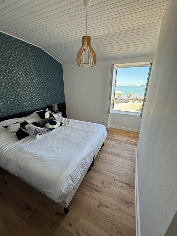 A bedroom with a bed, a hanging light fixture, a window with a view of the beach and a pier, and wooden flooring.