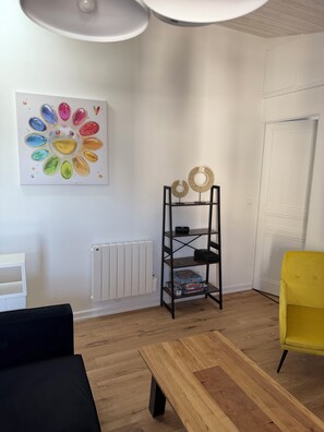 A room with a wooden floor, a black sofa, a yellow chair, a wooden coffee table, a radiator, a white door, and a shelf with decorative items.