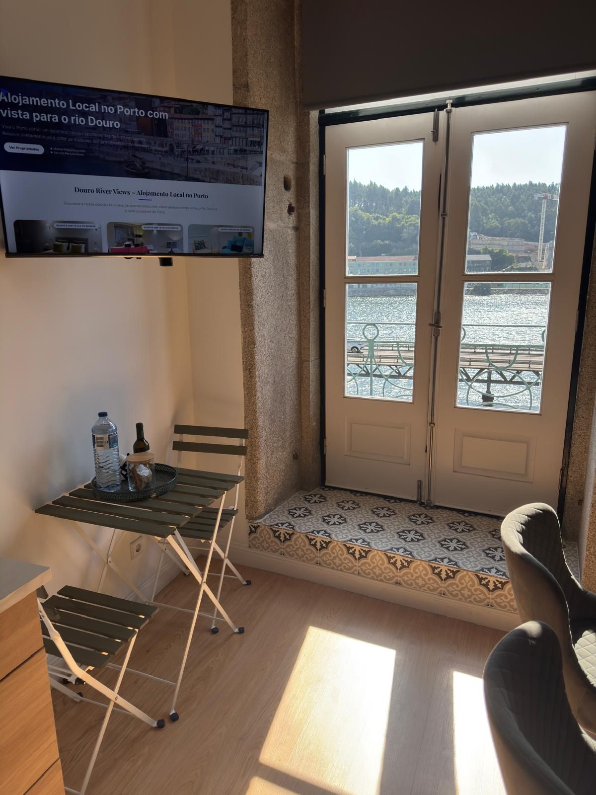Quarto com televisão na parede, uma mesa pequena com uma garrafa e uma garrafa de água, uma cadeira e vista para o rio através de uma porta de vidro.