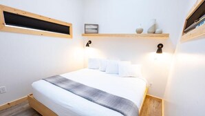 Un dormitorio con una cama de madera, ropa de cama blanca, una manta gris, un estante en la pared con objetos decorativos y una ventana con marco de madera.