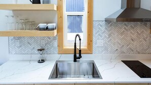 Una cocina con pileta de acero inoxidable, armarios de madera, un ventanal con persianas y un salpicadero con diseño en espiga.