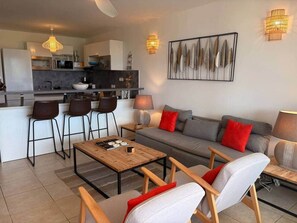 Uma sala de estar moderna com sofá, cadeiras, mesa de centro e cozinha ao fundo.
