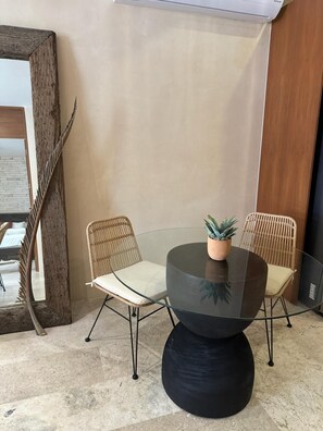 Un'area pranzo moderna con un tavolo in vetro e due sedie in vimini, una pianta in vaso sul tavolo e un grande specchio a sinistra.