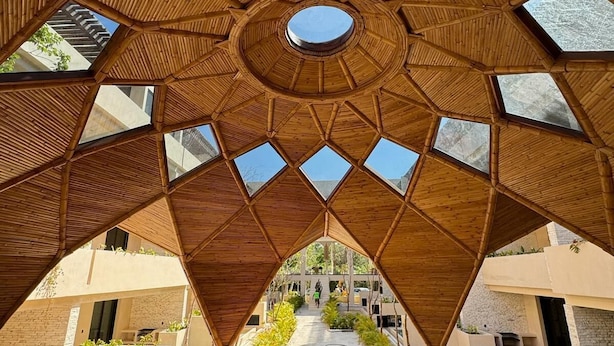 Una estructura de madera geométrica con varios tragaluces, que conduce a un espacio central abierto con vista a un edificio al fondo.