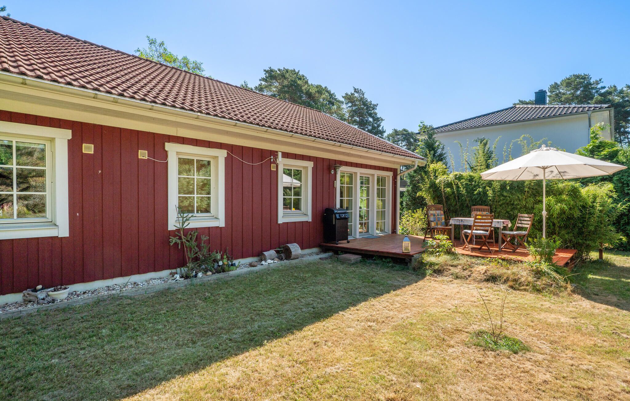 A red house with a tiled roof, a covered patio with a table and chairs, and a small garden area.