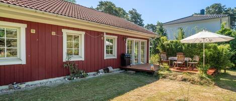 A red house with a tiled roof, a covered patio with a table and chairs, and a small garden area.