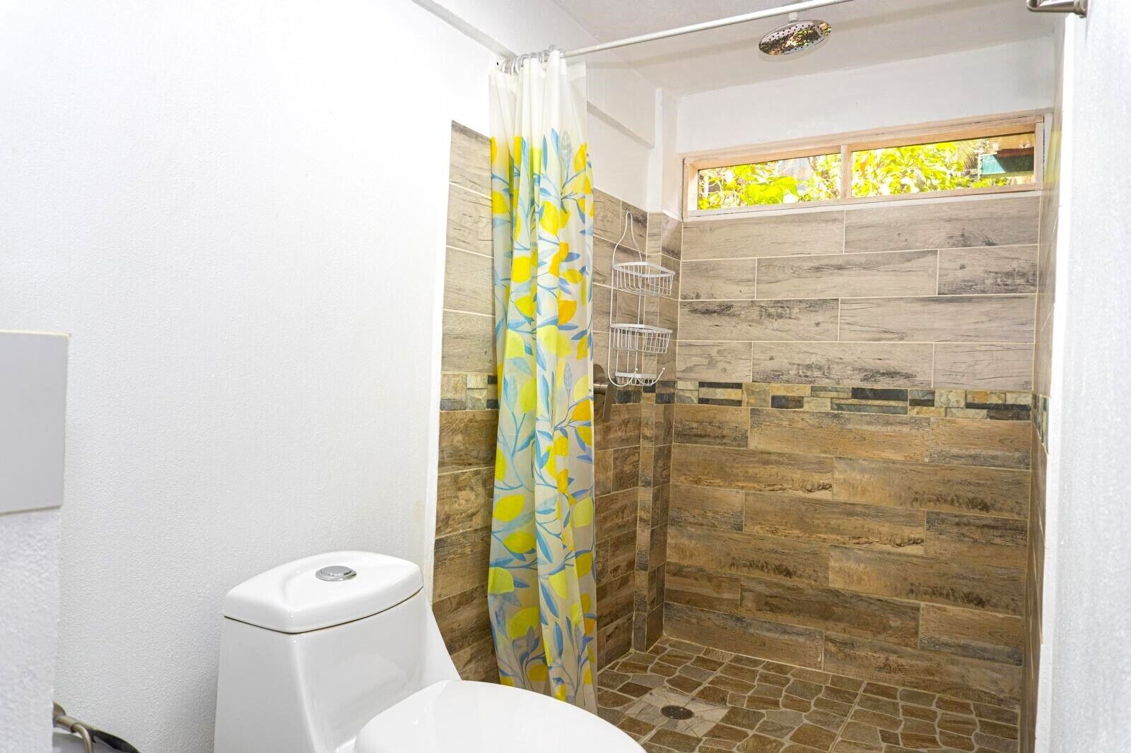 A bathroom with a shower, toilet, and a window with a view of greenery.