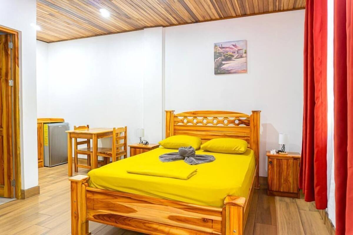Ein hölzernes Bett mit gelber Bettdecke, einem Holz-Kopfstück und zwei Kissen. Außerdem ein kleiner Holztisch mit Stuhl, ein Nachttisch mit Lampe und ein roter Vorhang.