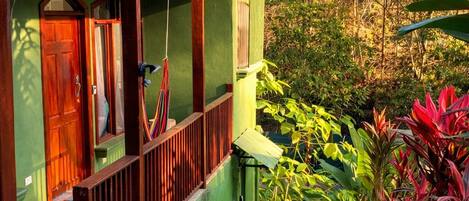 Ein grünes Gebäude mit einer Holztür, einer Veranda mit Geländer und einer Treppe, die in einen Garten mit verschiedenen Pflanzen führt.
