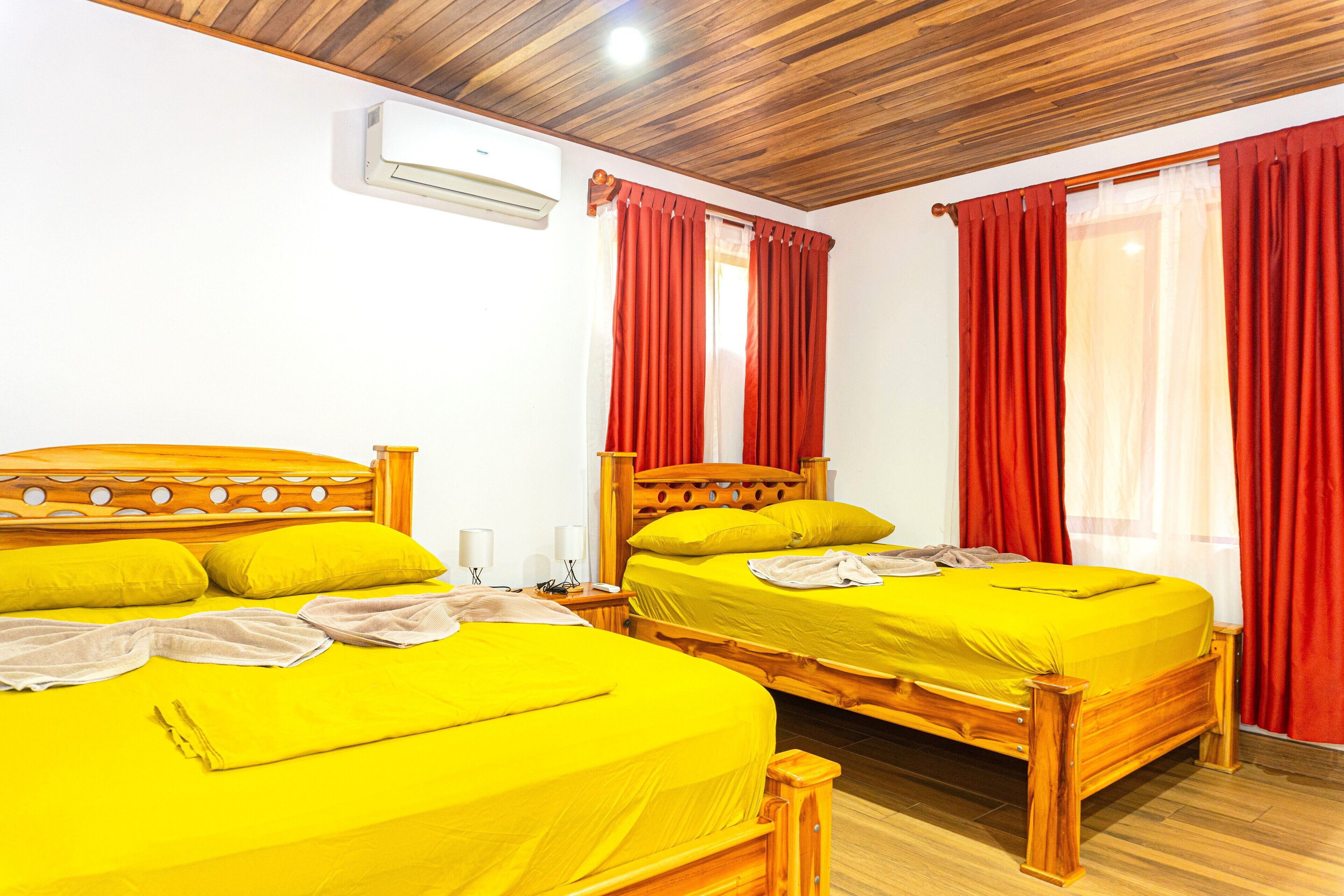 A room with two wooden beds, each with a yellow bedspread and matching pillows, red curtains, and an air conditioning unit on the wall.