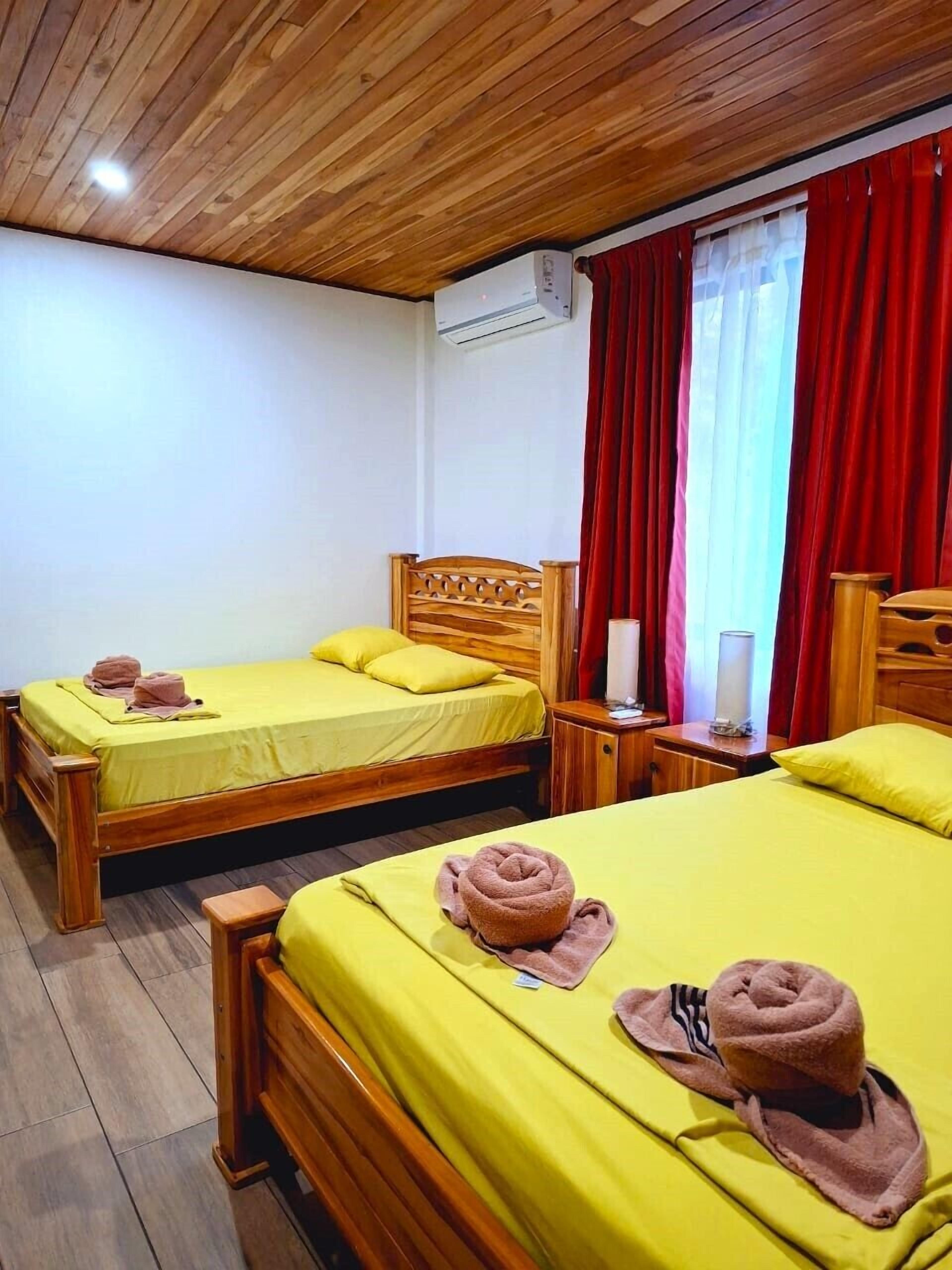 Une chambre d’hôtel avec deux lits en bois, une literie jaune, des rideaux rouges et un plafond en bois.