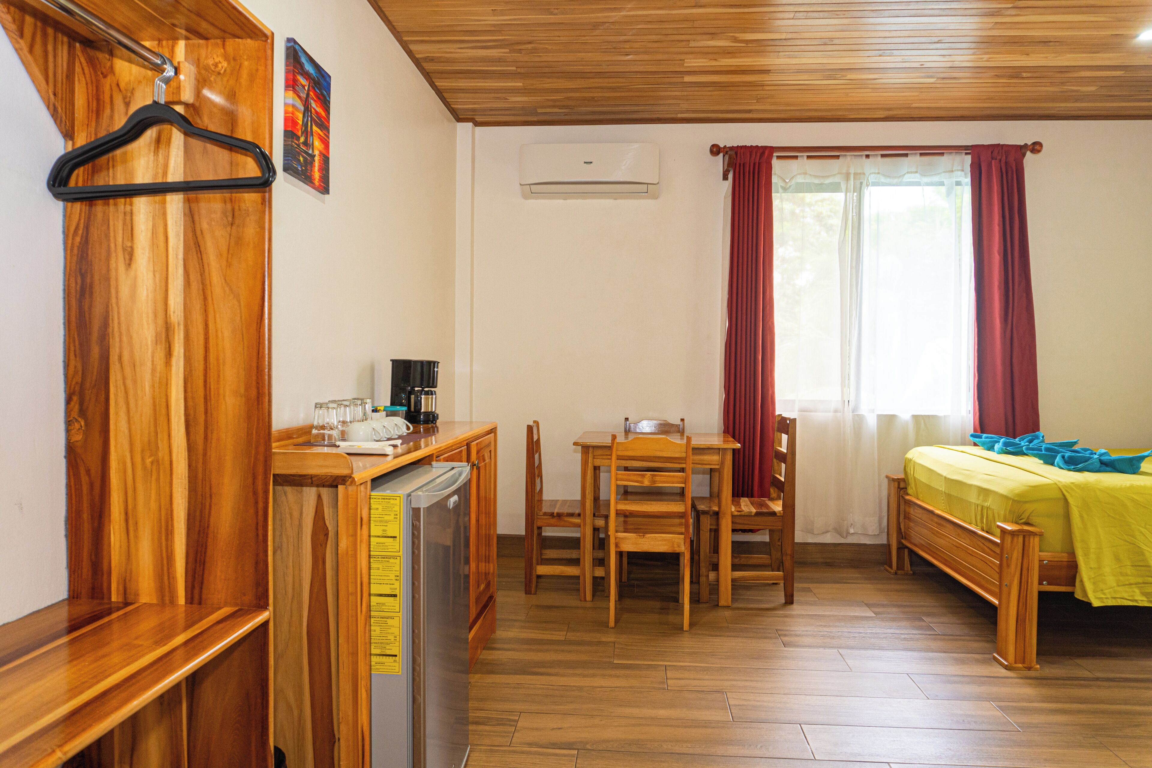 A compact room with a wooden floor, a bed with a yellow bedspread, a wooden table with chairs, a small kitchen area with a refrigerator, and a window with red curtains.