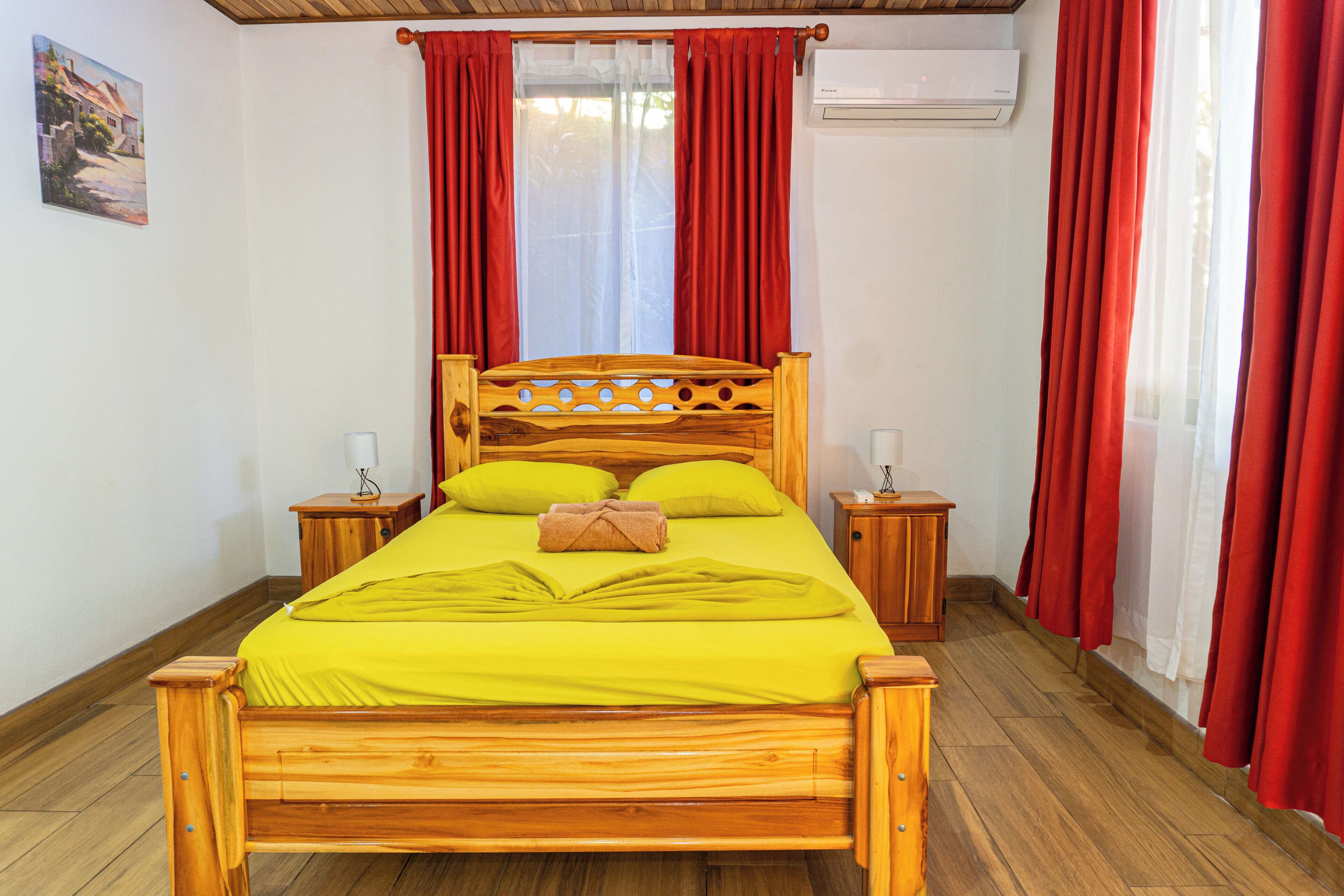 A wooden bed with a bright yellow bedspread, two bedside tables with lamps, red curtains, and an air conditioning unit on the wall.