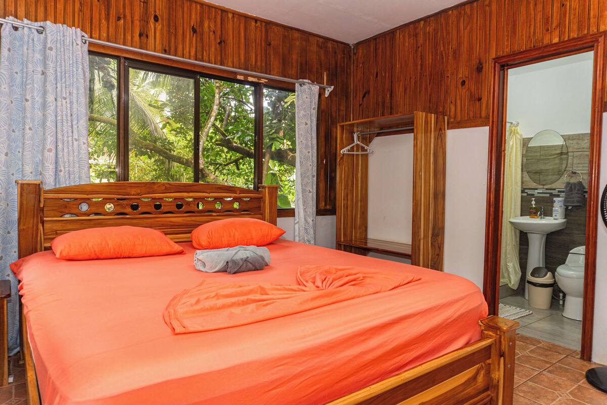 Ein Schlafzimmer mit Holzwänden, einem Bett, Vorhängen und Blick auf Grün.