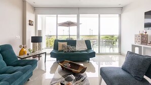 A modern living room with a teal sofa, a blue armchair, and a coffee table. There is a large window with a view of greenery outside. - First line beach in Cartagena edif.morros 3 (Cartagena)