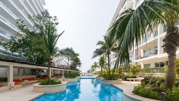A swimming pool with a tiled deck, surrounded by palm trees and plants, with modern buildings on either side. - First line beach in Cartagena edif.morros 3 (Cartagena)