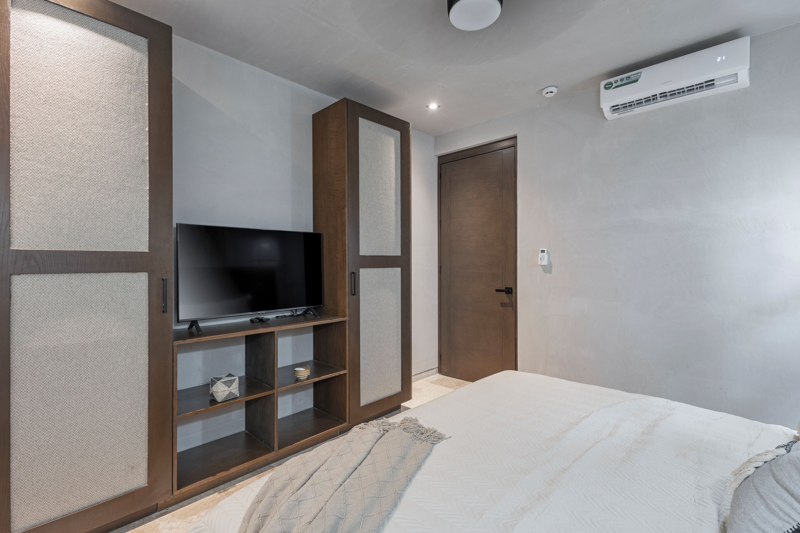 A bedroom with a bed, a TV on a wooden cabinet, a wardrobe, and an air conditioning unit on the wall.