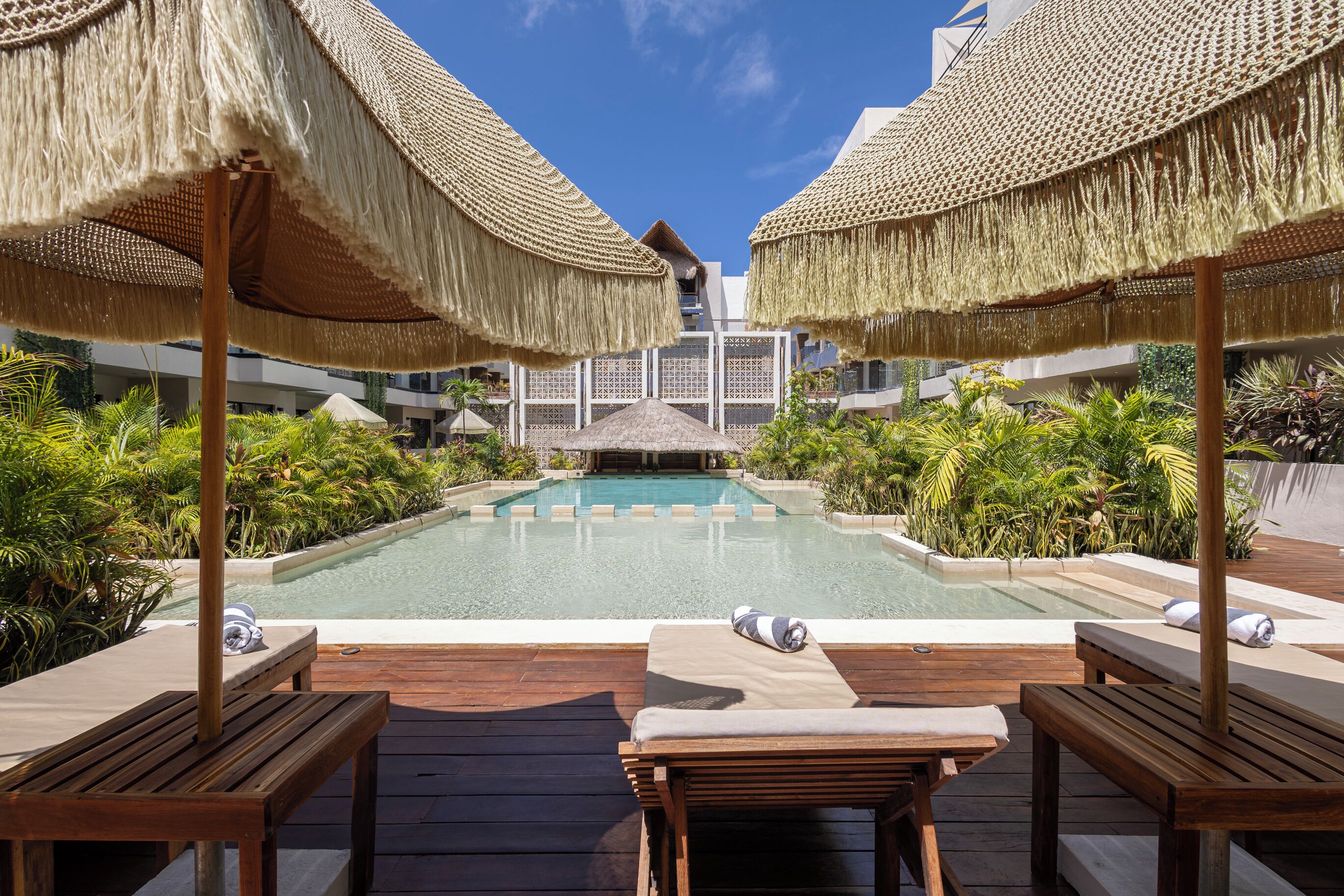 Un espace piscine agrémenté de chaises longues en bois et de parasols en chaume, le tout entouré d’une végétation luxuriante.