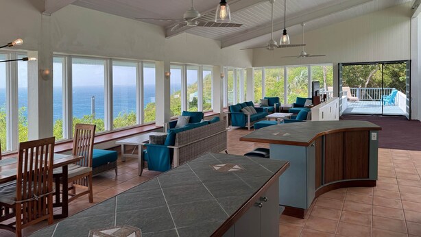 A spacious room with a large window overlooking the ocean, a seating area with blue furniture, and a kitchen counter. - H2O Eco Villa (ST Thomas)